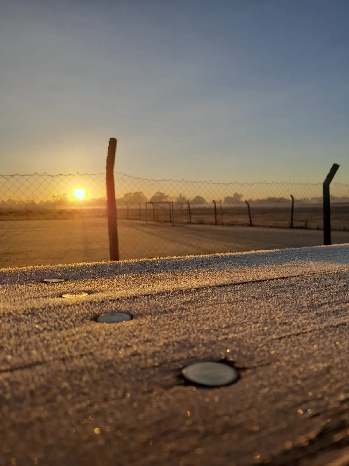 Tablón de tribuna cubierto de helada al amanecer, con una cancha de fútbol de fondo